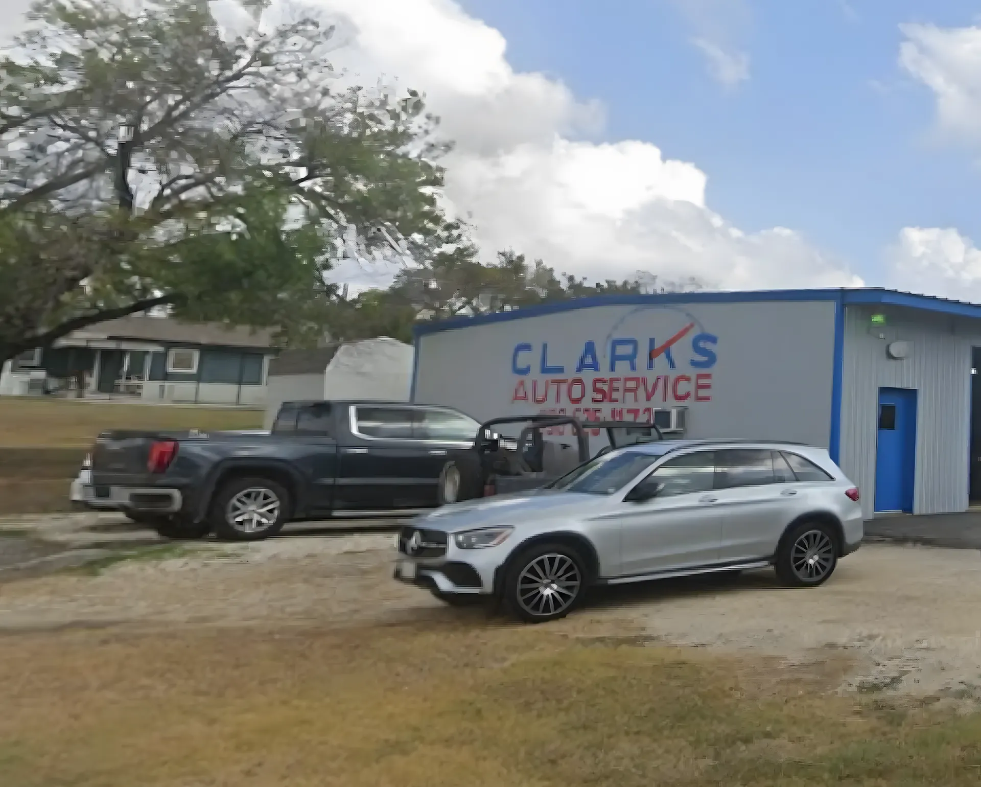 About us, auto repair in New Braunfels, TX by Clark's Auto Service. A small auto service shop with a sign reading CLARKS AUTO SERVICE; a black pickup truck and a silver SUV are parked outside on a grassy lot, with trees and a house in the background.