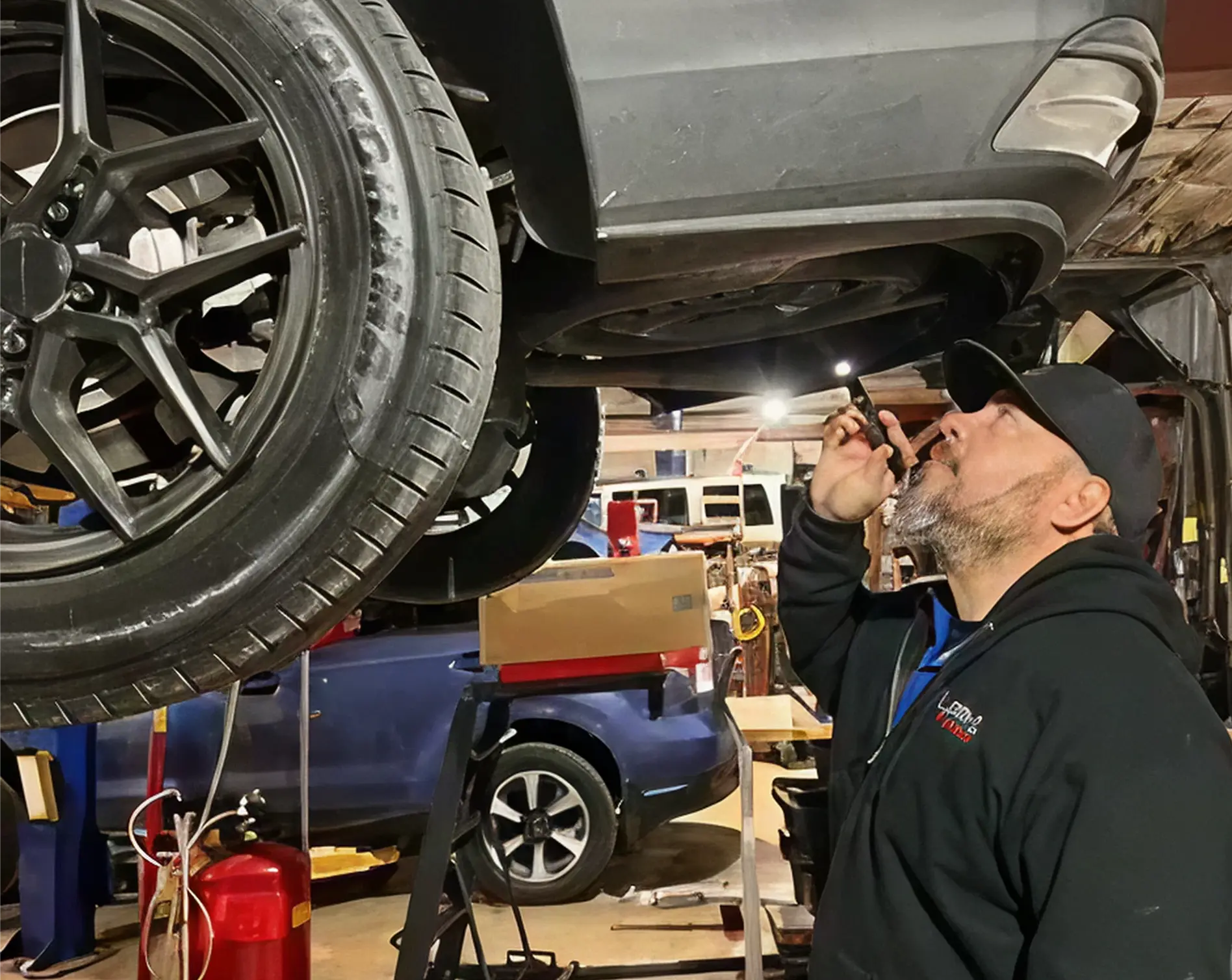 Auto repair in New Braunfels, TX by Clark's Auto Service. A mechanic wearing a black cap and jacket inspects the underside of a car raised on a lift in a garage, holding a flashlight and looking up at the vehicle. Tools and other cars are visible in the background.