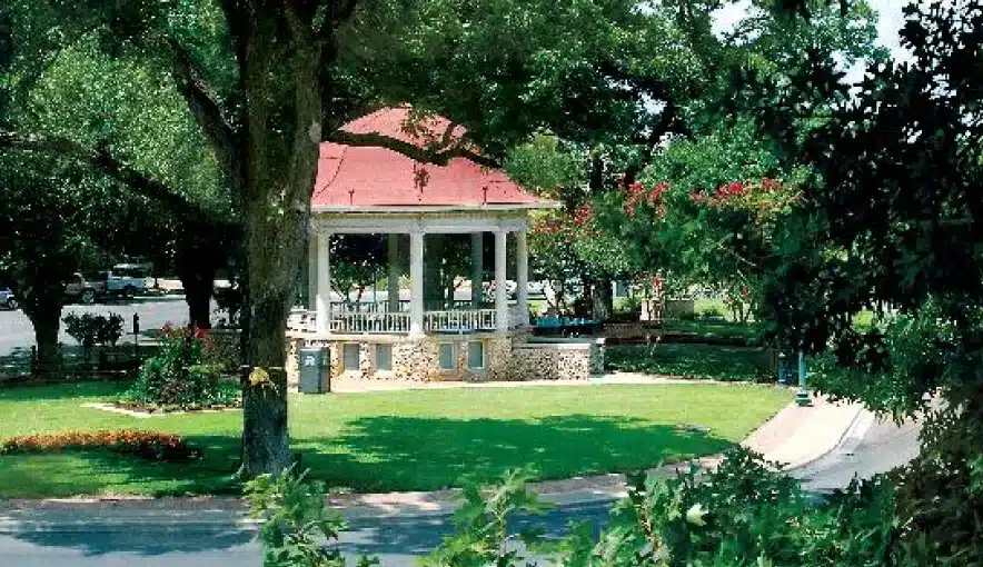 A white gazebo with a red roof stands in the middle of a green park, surrounded by trees and landscaped grass, with a parking lot visible in the background.