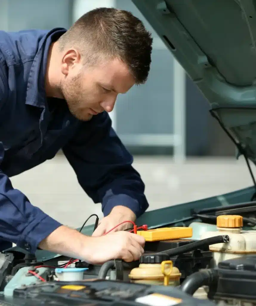 A mechanic wearing a blue uniform inspects a car engine with the hood open, using diagnostic cables and focusing intently on the vehicle’s components.