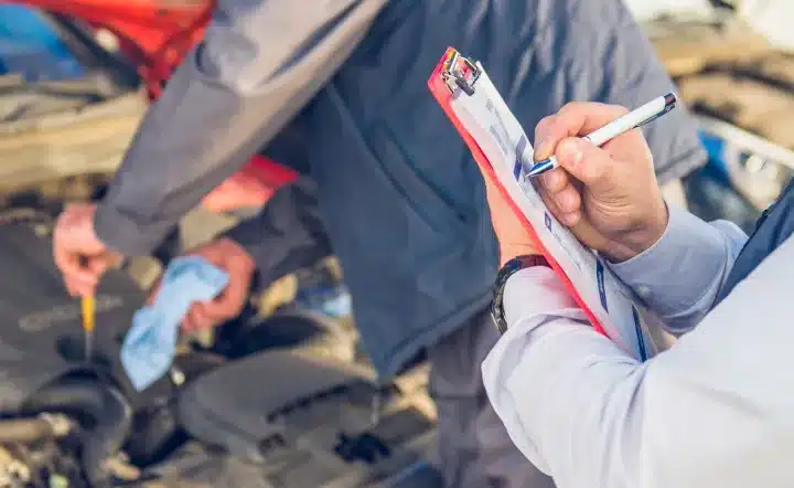 A person holds a clipboard and writes while another person checks a car engine, suggesting a vehicle inspection or maintenance process.
