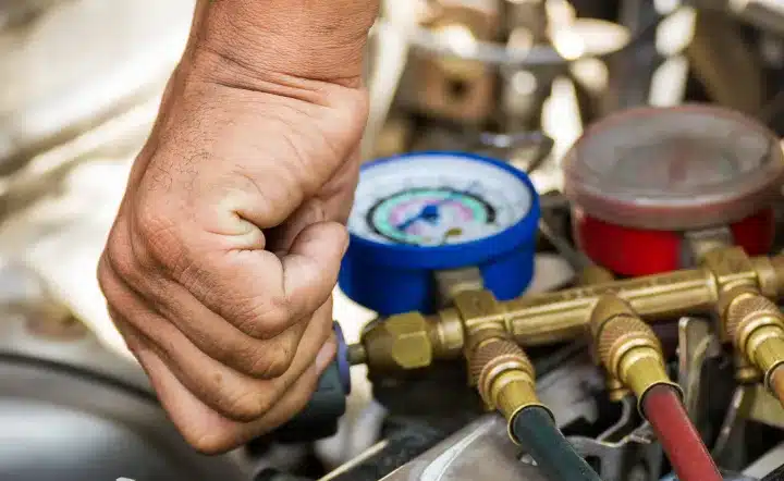 A close-up of a persons hand adjusting a gauge on an HVAC manifold with red, blue, and yellow hoses, likely during air conditioning maintenance or repair.