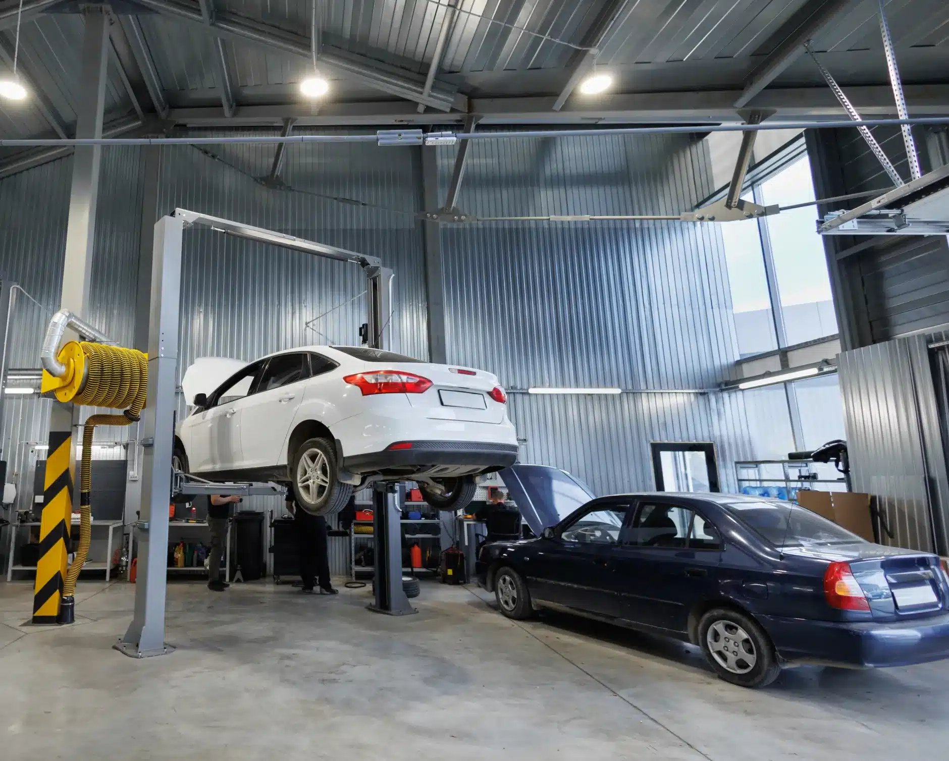 Auto repair in New Braunfels, TX by Clark's Auto Service. A white car is elevated on a hydraulic lift in a modern auto repair shop, while a dark sedan is parked nearby with its hood open. The workshop features large windows and metal walls.