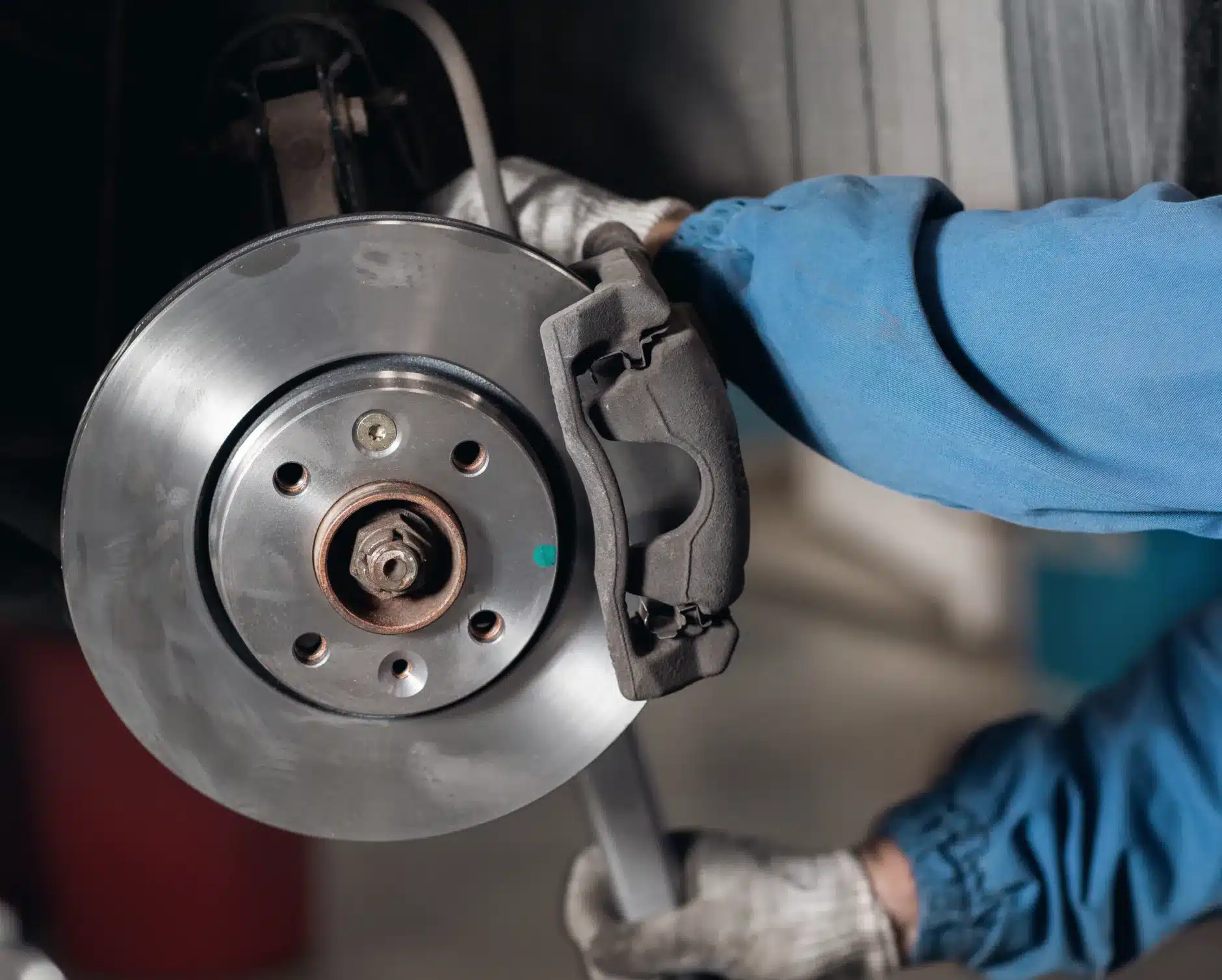 Brake repair, auto repair in New Braunfels, TX by Clark's Auto ServiceA person wearing gloves and a blue uniform is replacing or inspecting a car brake disc and caliper, using a tool to work on the vehicles braking system.