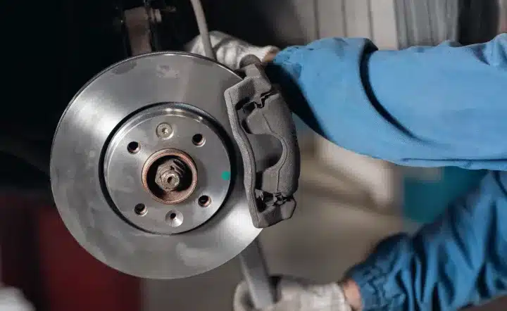 A mechanic wearing gloves and a blue sleeve works on a car’s exposed brake disc and caliper, performing maintenance or repair inside a garage.