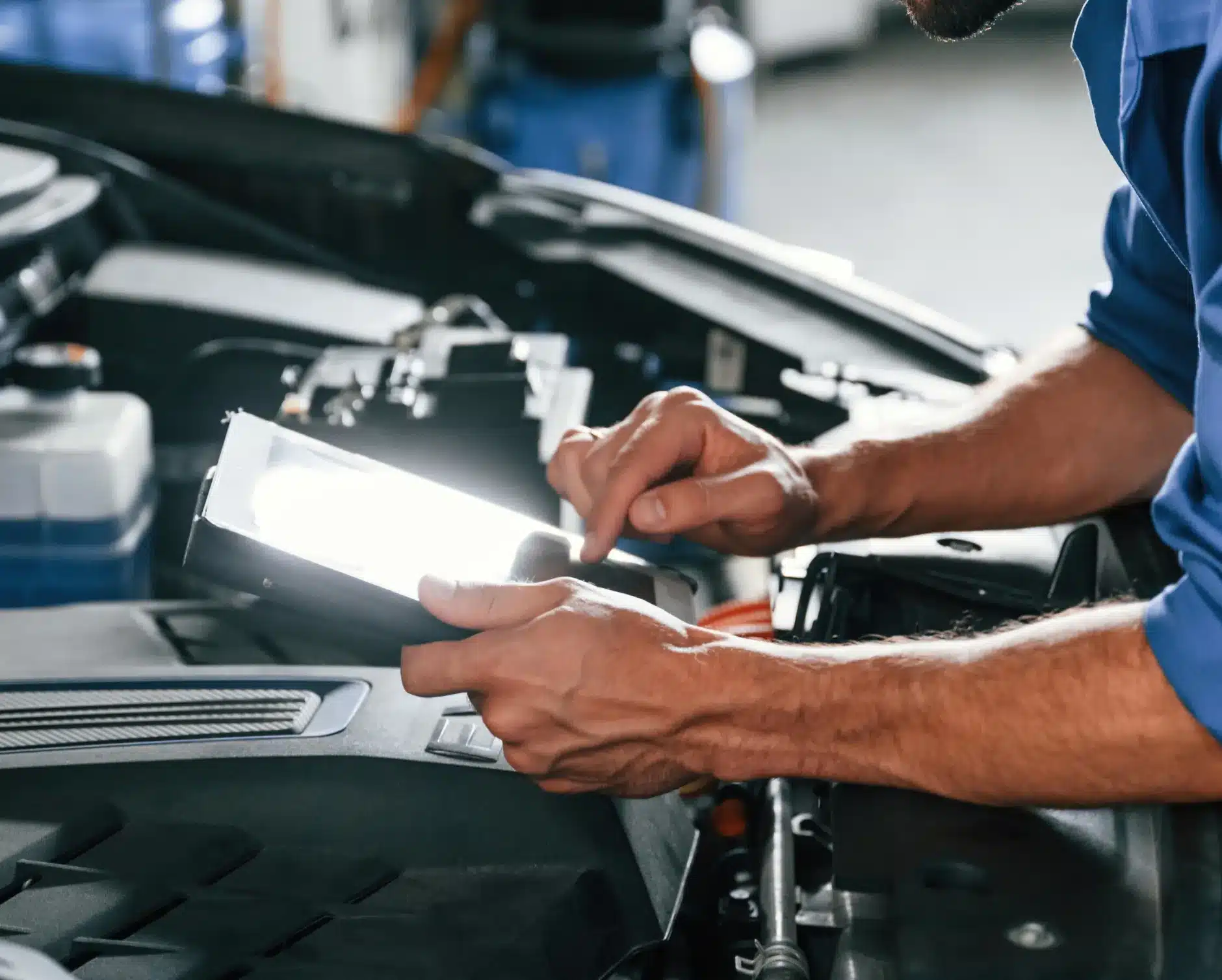 Car diagnostics, auto repair in New Braunfels, TX by Clark's Auto Service. A mechanic in a blue uniform uses a digital tablet while inspecting the engine of a car, with the vehicles hood open and various engine components visible.