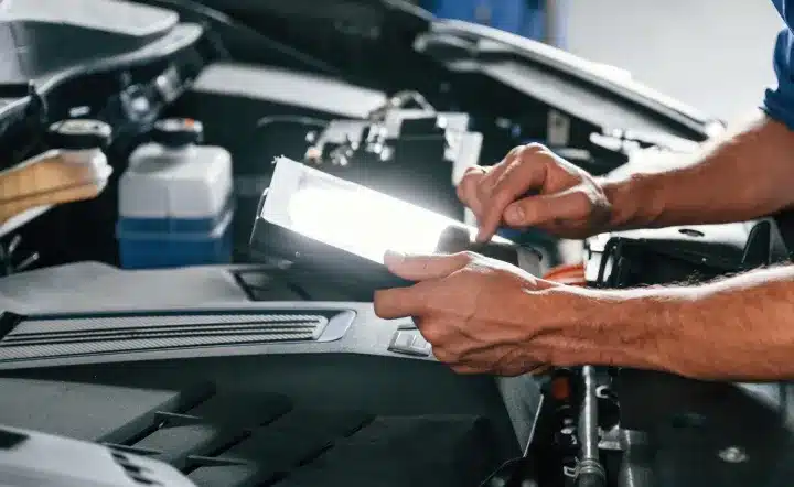 A person using a digital tablet to inspect or diagnose a car engine in a garage or workshop. The car hood is open, revealing engine components.