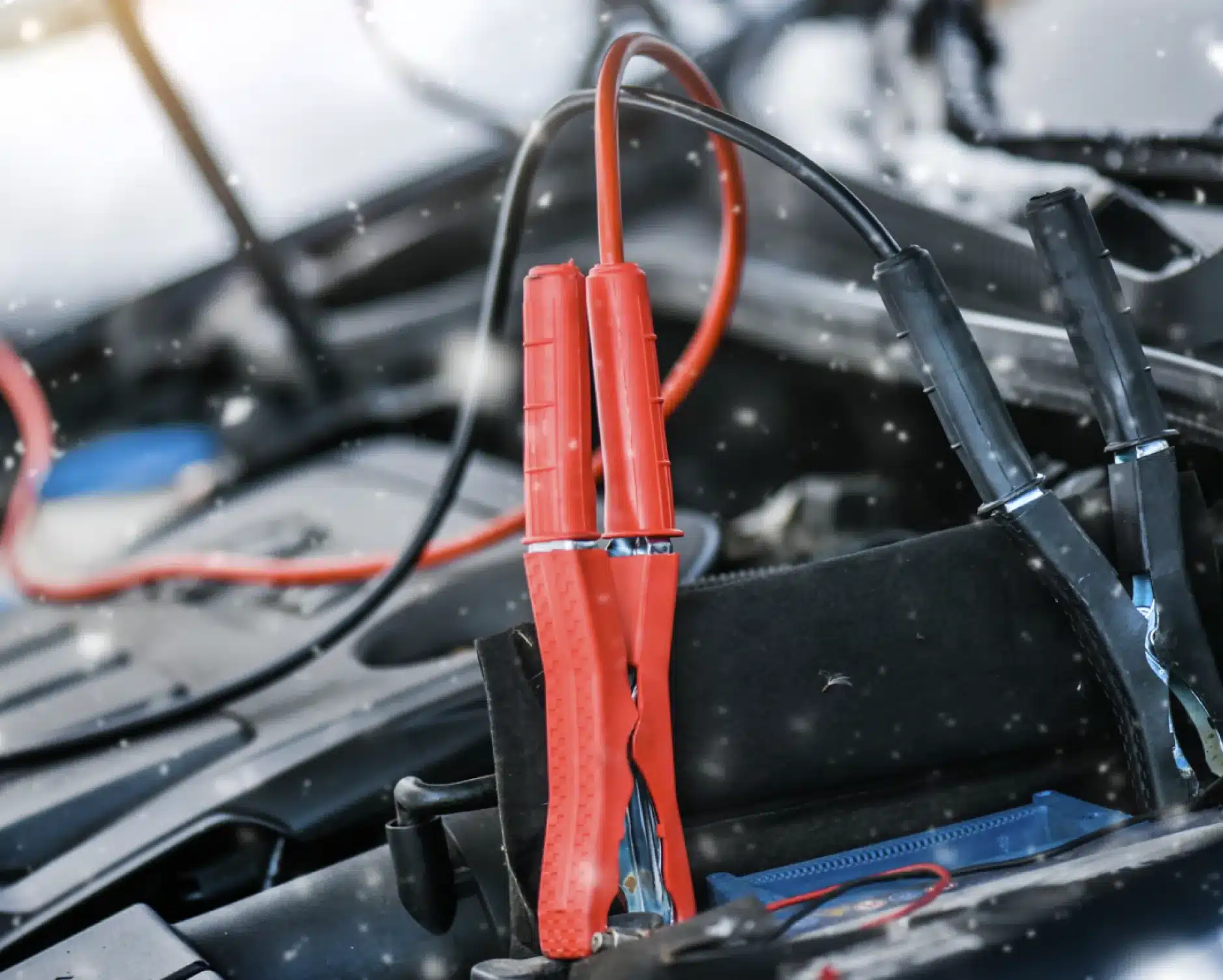 Auto electrical repair, auto repair in New Braunfels, TX by Clark's Auto Service. Close-up of red and black jumper cables attached to a car battery, with a blurred car engine in the background, indicating a vehicle jump-start in progress.