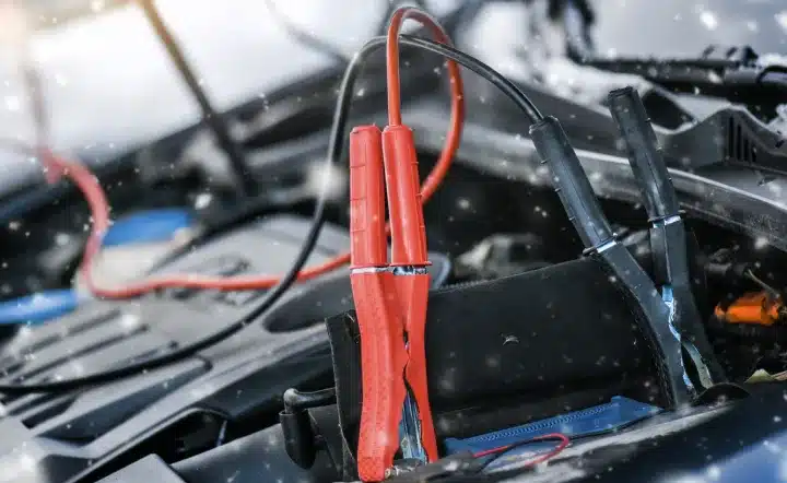Close-up of red and black jumper cables attached to a car battery under an open hood, with some snow gently falling on the engine components.