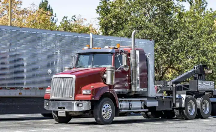 A red semi-truck cab is parked on a street in front of a large silver trailer, with trees and bright sunlight in the background.