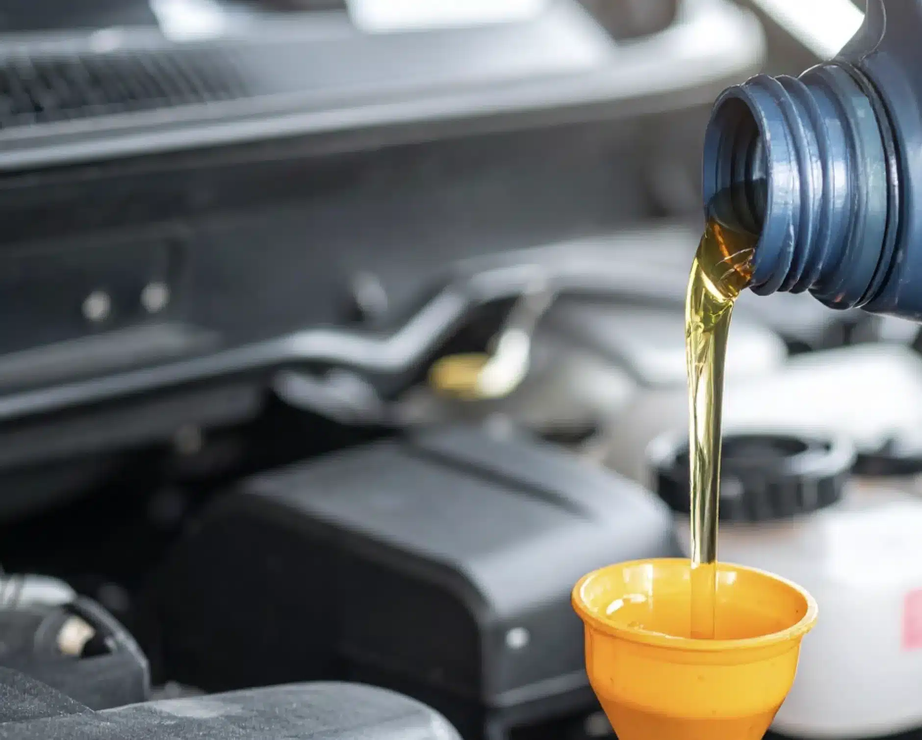 Motor oil being poured from a blue bottle into a yellow funnel, with a blurred car engine in the background.