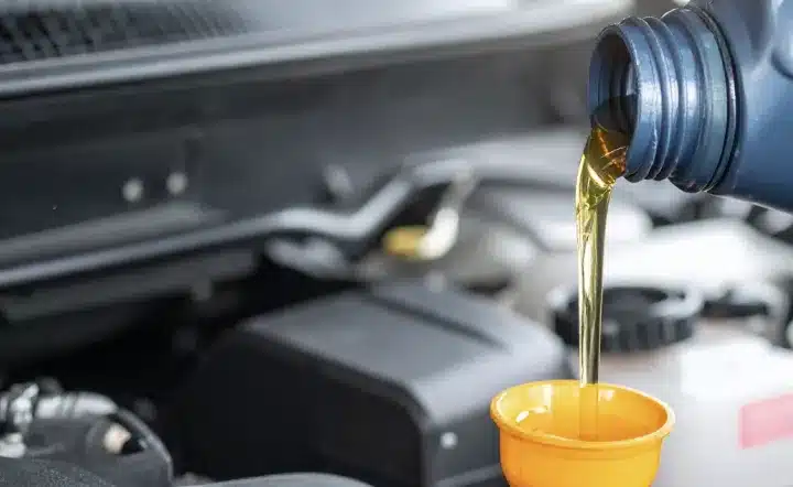 Close-up of motor oil being poured from a blue bottle into a yellow funnel under the hood of a car, with engine components visible in the background.