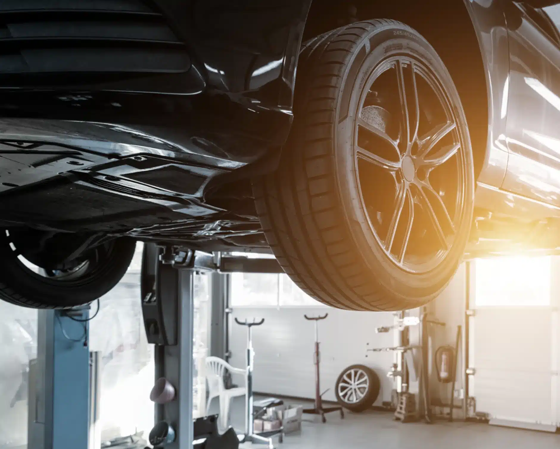 Preventative maintenance, auto repair in New Braunfels, TX by Clark's Auto Service. A close-up view of a car’s rear wheel while the vehicle is lifted on a hydraulic lift inside a brightly lit auto repair garage. Sunlight shines through a window in the background.