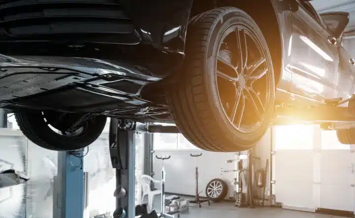 A black car is lifted on a hydraulic platform in a well-lit garage, with sunlight streaming through the window and another tire visible in the background.