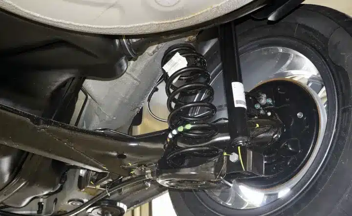 Suspension repair, auto repair in New Braunfels, TX by Clark's Auto Service. Close-up view of a car’s rear suspension system, showing a coil spring, shock absorber, axle, and part of the wheel with a visible brake assembly underneath the vehicle.