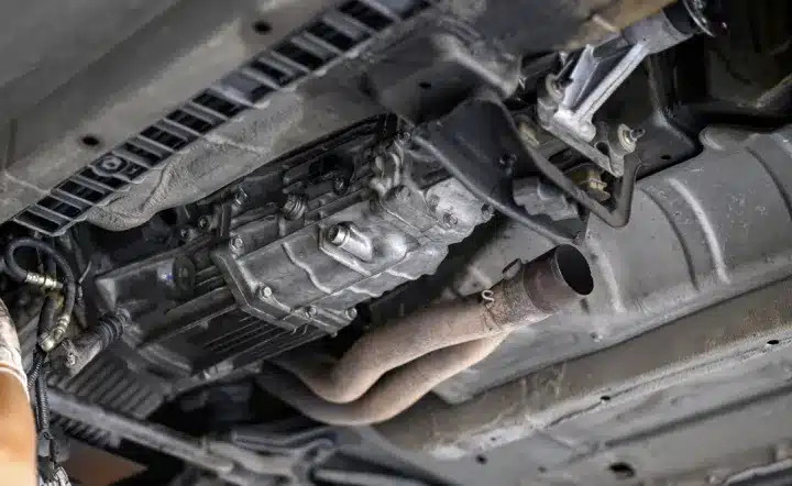 A close-up view of the underside of a vehicle, showing the exhaust pipe, transmission, and various mechanical components. A persons gloved hand is visible to the left, appearing to work on the car.