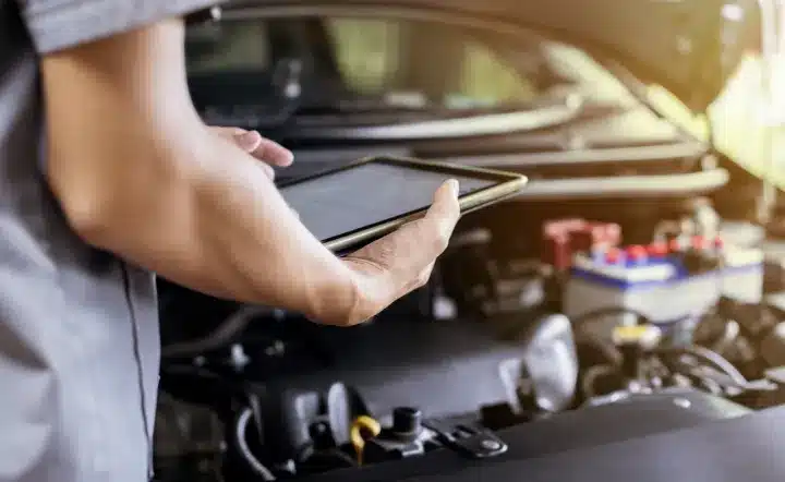 A person holds a tablet while standing next to an open car hood, possibly diagnosing or inspecting the vehicle’s engine using digital tools. The focus is on the arm, tablet, and car engine.
