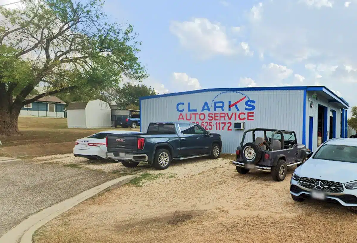 Small auto repair shop called Clarks Auto Service with several vehicles, including trucks, a Jeep, and a white sedan, parked outside on a gravel lot. The shop is a blue and white metal building.